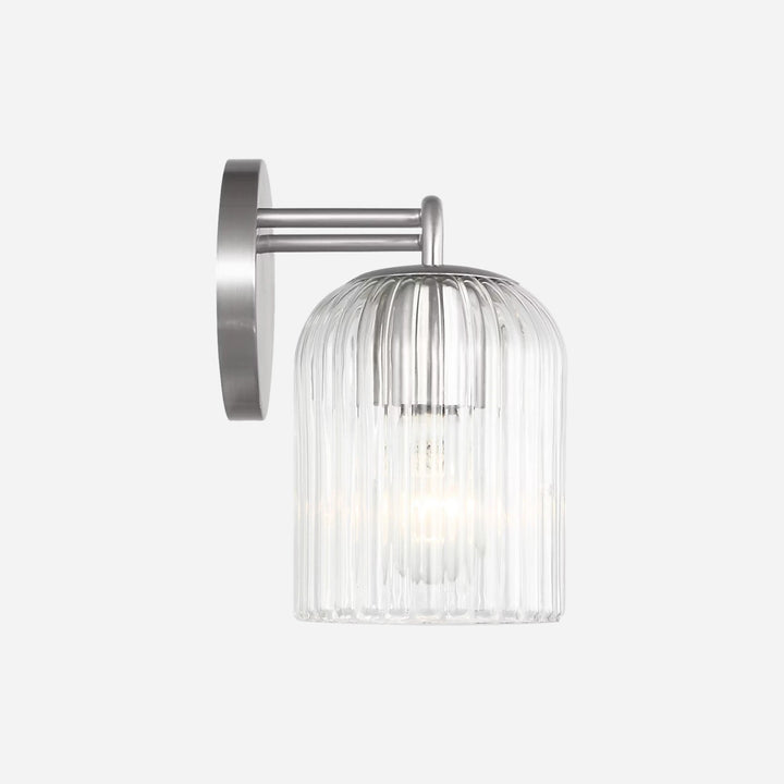 Applique murale Maison Olive Eisele avec support en métal brossé et abat-jour cylindrique en verre strié transparent.