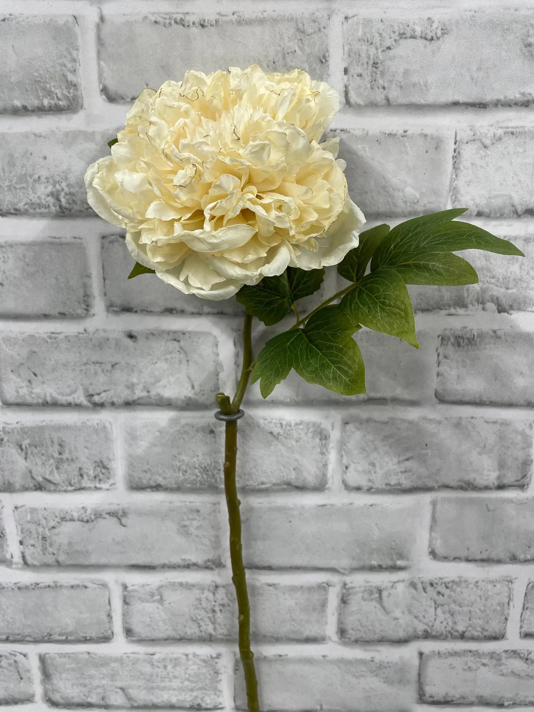 Grande pivoine artificielle Maison Olive couleur crème avec pétales détaillés et feuilles vertes sur fond brique