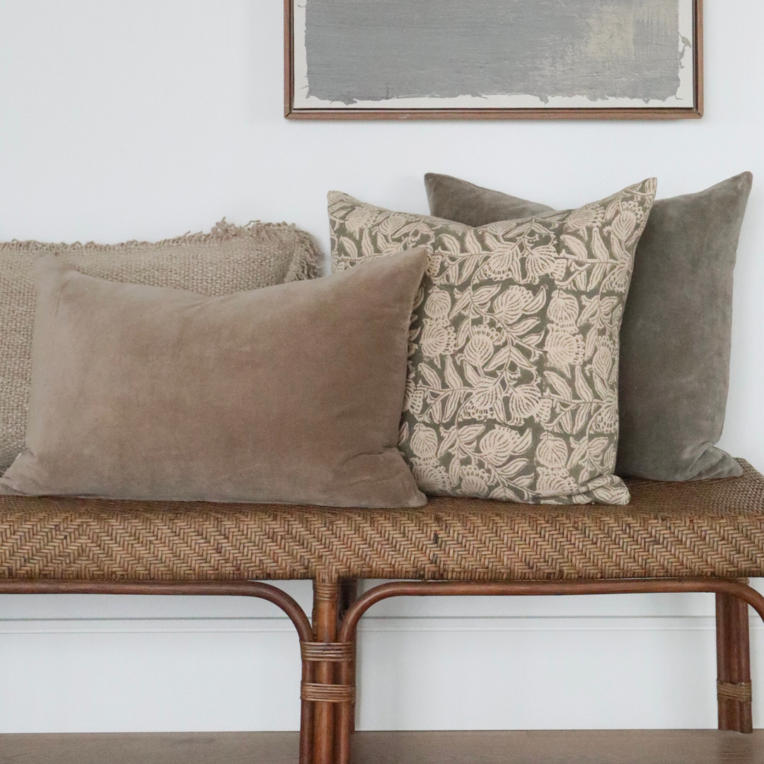 Housse de coussin Maison Olive en coton beige et gris à motif floral artisanal posée sur un banc en rotin