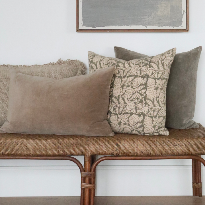 Housse de coussin Maison Olive en coton beige et gris à motif floral artisanal posée sur un banc en rotin