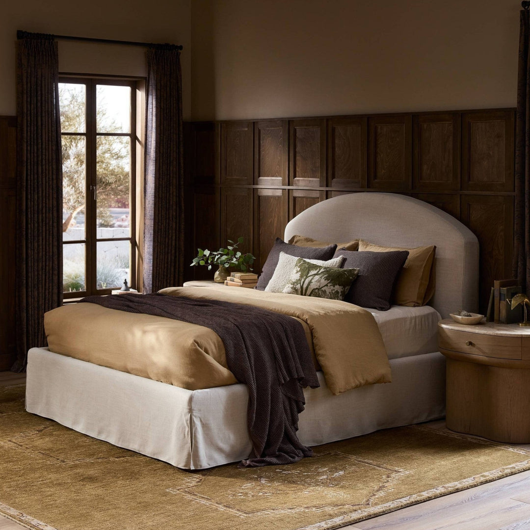 Lit Maison Olive capitonné crème avec jupe et tête de lit arquée, drapé d'une couverture marron et de coussins décoratifs.