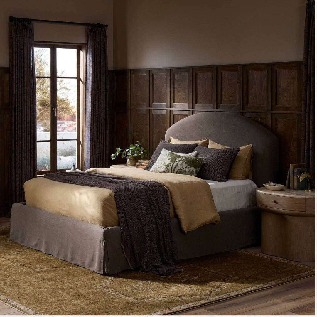 Lit capitonné Maison Olive avec jupe et tête de lit arquée en tissu texturé crème, literie dorée et couverture marron.