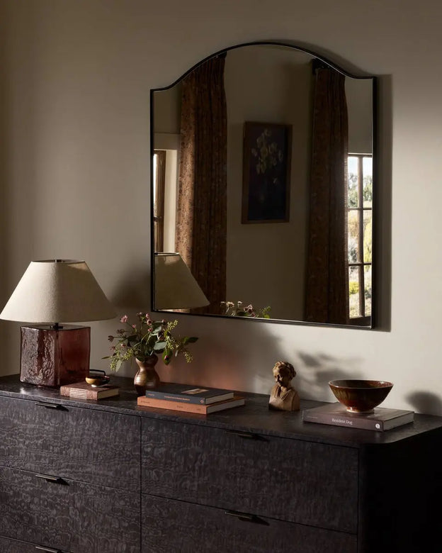Miroir arqué noir au-dessus d’une commode décorée d’une lampe et d’objets