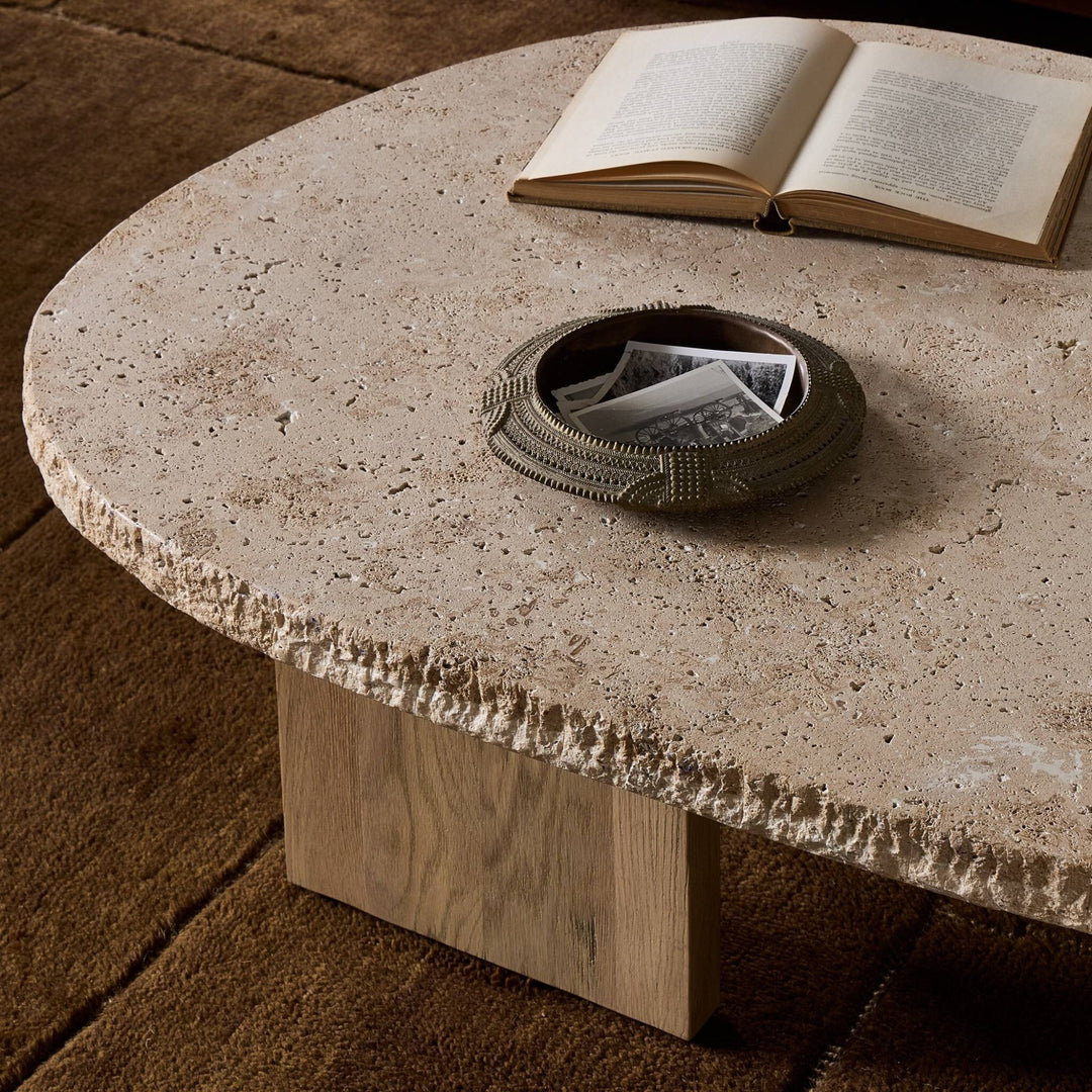 Maison Olive table basse ovale en travertin beige texturé avec un livre ouvert et un vide-poche décoratif.