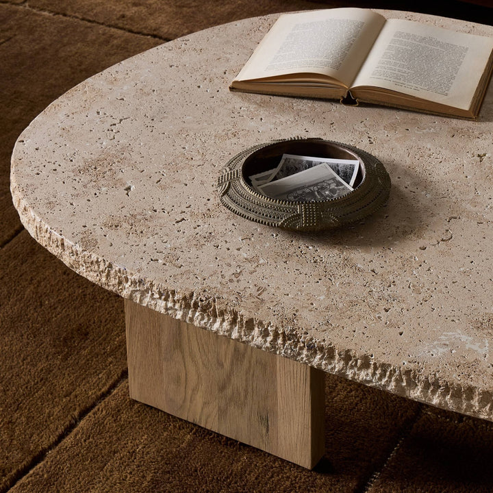 Maison Olive table basse ovale en travertin beige texturé avec un livre ouvert et un vide-poche décoratif.