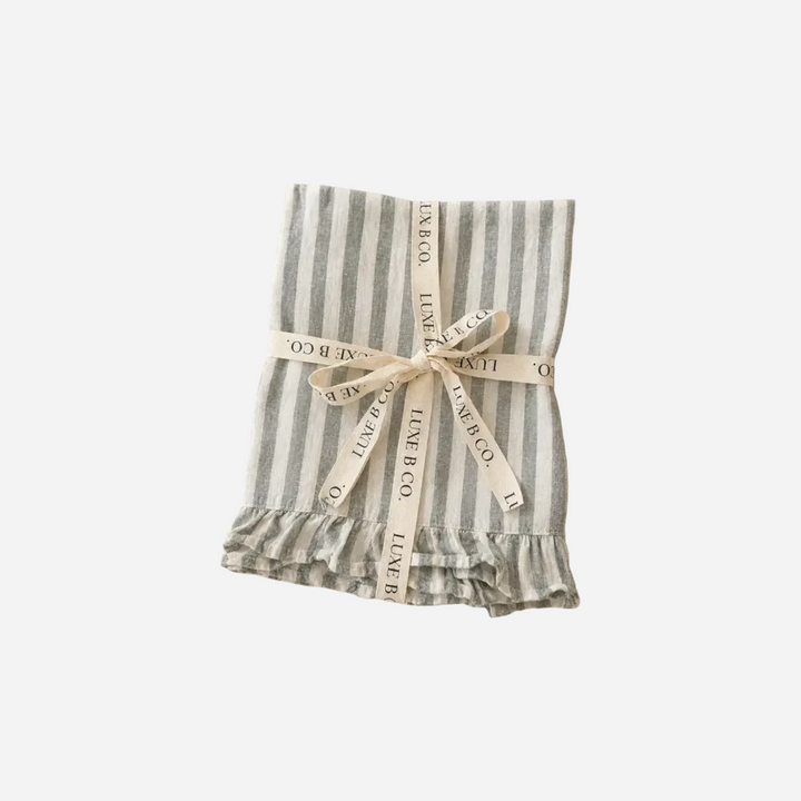 Torchon de cuisine Maison Olive en lin rayé gris et blanc avec finition à volants, noué par un ruban décoratif.